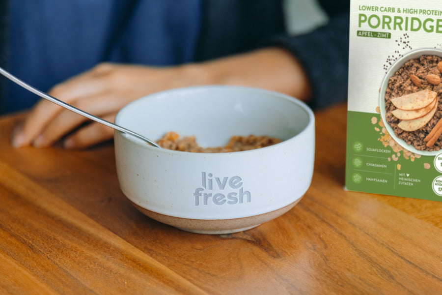 Schüssel mit der Aufschrift „Live Fresh“, die eine Portion Porridge enthält. Neben der Schüssel steht eine Packung „Live Fresh Porridge Apfel-Zimt“, die die Zutaten Sojaflocken, Chiasamen und Hanfsamen hervorhebt. Eine Hand ist im Hintergrund zu sehen, während ein Löffel im Porridge ruht, was auf eine gesunde und nahrhafte Mahlzeit hinweist.