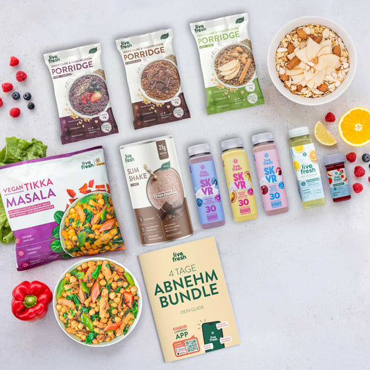 Flatlay verschiedener LiveFresh Produkte auf hellem Hintergrund mit Porridge Beuteln, Slim Shake, SKVR Protein Drinks, Saft Shots und veganer Tikka Masala Mahlzeit neben frischem Obst und einer Schüssel Essen. In der Mitte liegt ein Heft mit der Aufschrift 4 Tage Abnehm Bundle Dein Guide.