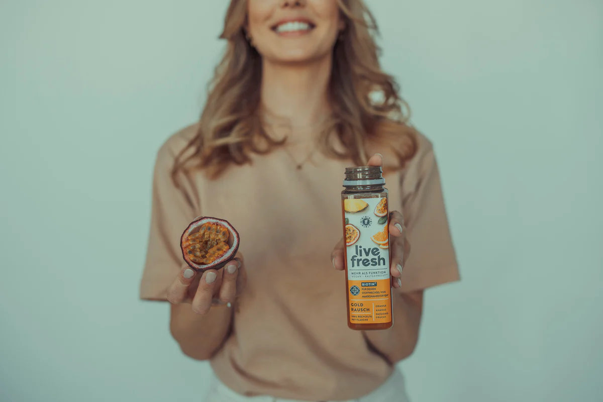 Eine lächelnde Frau in einem beigefarbenen Shirt hält in einer Hand eine halbierte Maracuja und in der anderen eine geöffnete Flasche LiveFresh Saft „Goldrausch“ mit Orange, Mango und Maracuja. Der Hintergrund ist ein grau-grünlicher Farbton.
