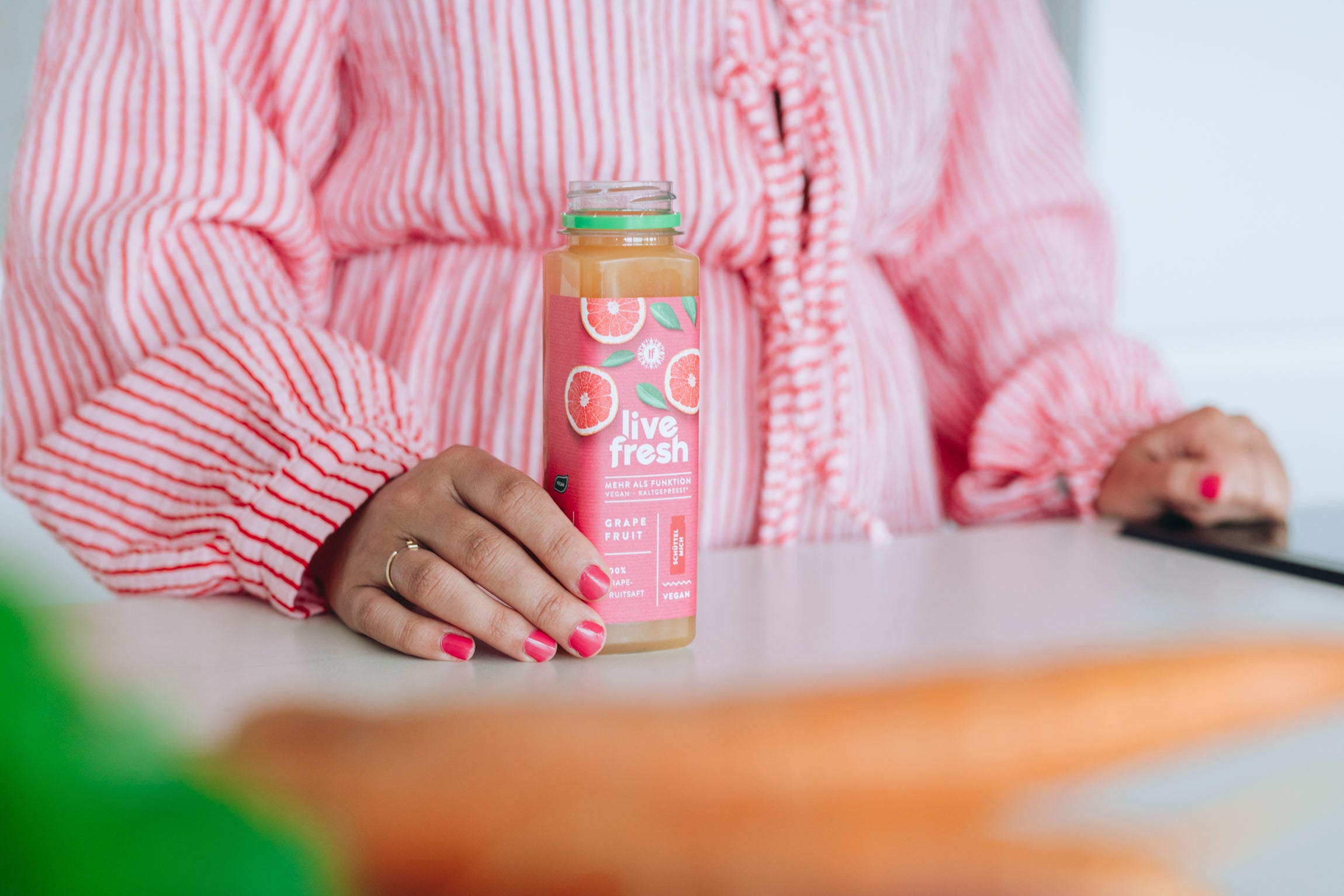 Frau in rosa-weiß gestreifter Bluse steht an einem Tisch und hält eine Flasche „Live Fresh“ Grapefruitsaft mit pinkem Etikett.