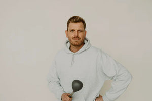 Ein Mann mit kurzem Bart und hellem Hoodie steht vor einem neutralen Hintergrund. Er hält eine schwarze Suppenkelle in der Hand und blickt selbstbewusst in die Kamera.