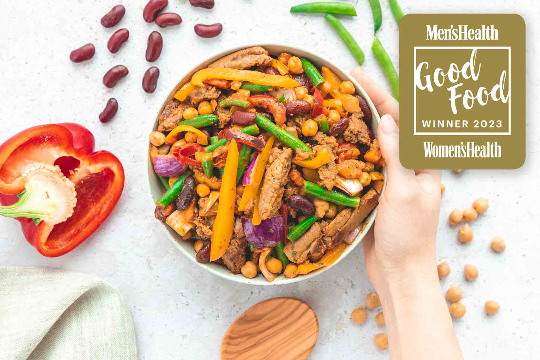Eine Hand hält eine Schüssel auf einer hellen Oberfläche mit einer Mahlzeit aus pflanzlichem Protein, Kichererbsen, grünen Bohnen, Kidneybohnen, Paprika und roten Zwiebeln. Daneben eine halbierte Paprika, Bohnen und Kichererbsen. Oben rechts ein goldenes Siegel mit der Aufschrift „Men’sHealth & Women’sHealth Good Food Winner 2023“.