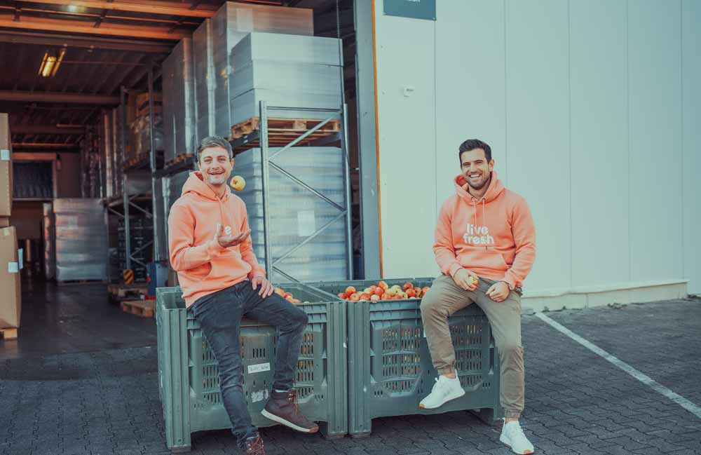 Zwei junge Männer in orangenen "LiveFresh"-Hoodies sitzen lachend auf einer großen grünen Obstkiste vor einem Lagerhaus. Einer von ihnen wirft einen Apfel in die Luft, während der andere eine Flasche hält. Die Kiste ist mit frischem Obst gefüllt, und im Hintergrund sind Paletten und Lagerregale sichtbar.