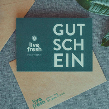 Dunkelgrüner Gutschein von LiveFresh mit weißer Aufschrift GUTSCHEIN und Logo auf einer beigen Briefumschlaghülle, platziert auf einem grauen Stoffuntergrund mit grünen Blättern am Rand.