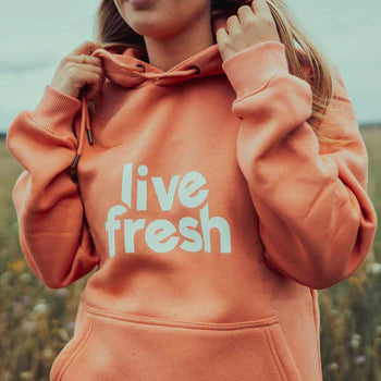 Person mit blonden Haaren in einem orangefarbenen Hoodie mit weißem Livefresh-Schriftzug, die leicht an der Kapuze zieht. Hintergrund mit unscharfer Naturkulisse.