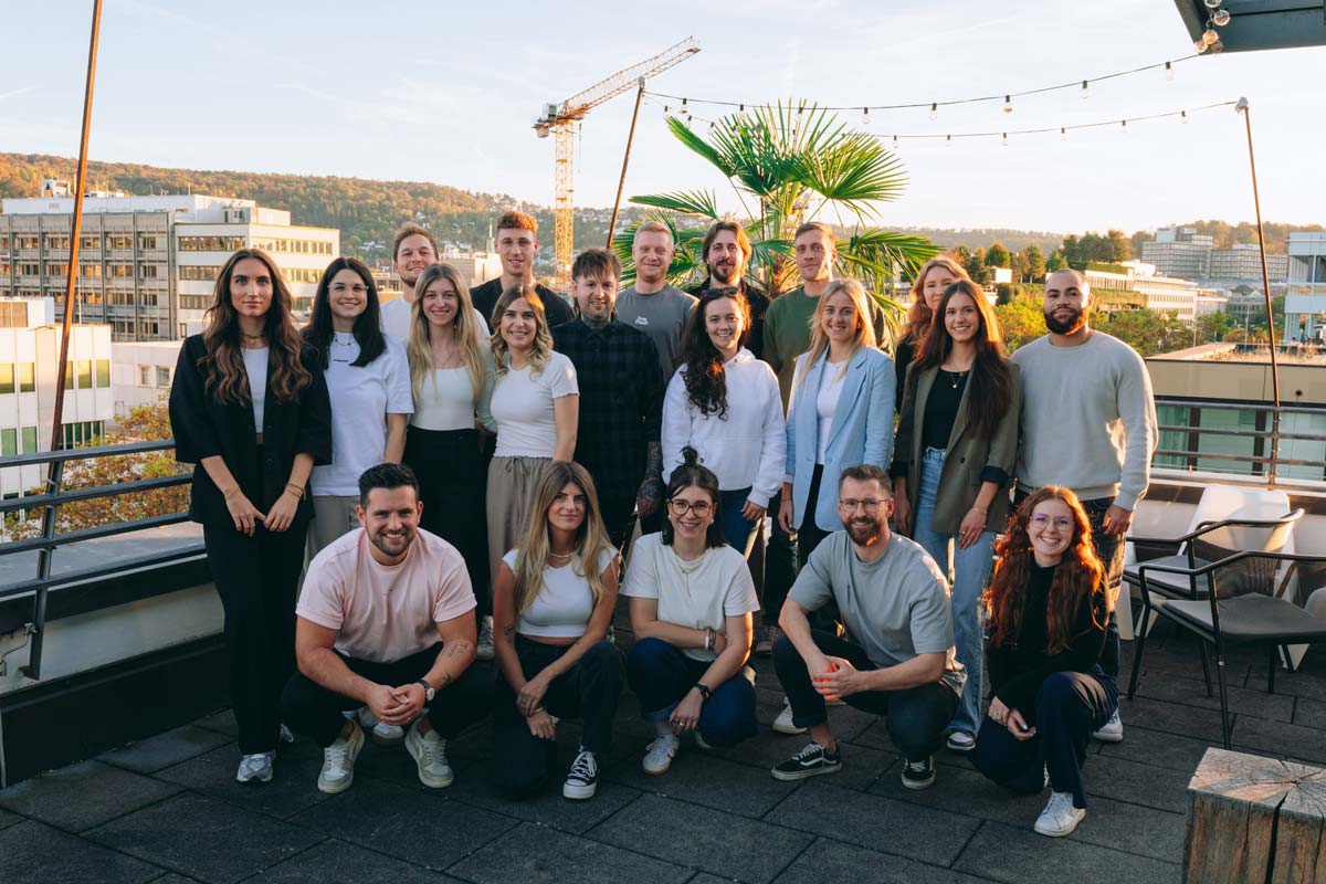 Gruppe von Menschen auf einer Dachterrasse bei Sonnenuntergang, mit Blick auf eine Stadtlandschaft mit Gebäuden, einem Kran und einer Palme. Einige Personen hocken sich hin, die anderen stehen. Alle lächeln und posieren für ein gemeinsames Foto.