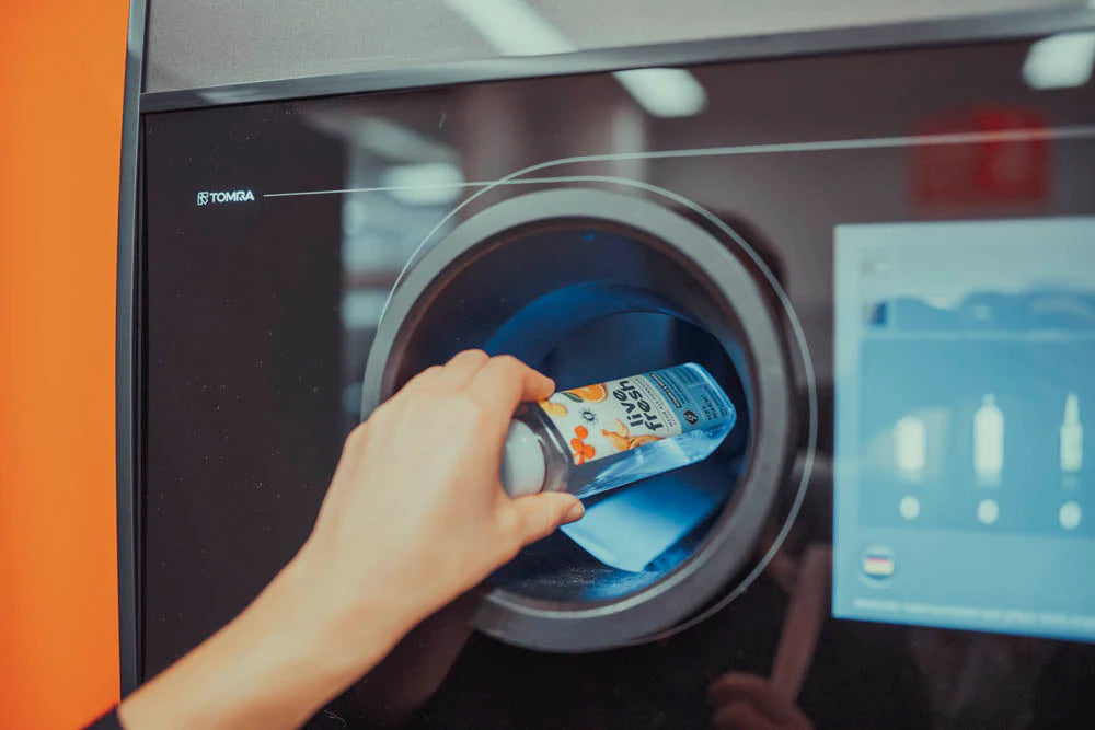 Eine Hand, die eine leere Live Fresh Saftflasche in einen Pfandautomaten einwirft. Die Flasche wird in den runden Eingabeschacht der Maschine eingeführt, während ein Bildschirm auf der rechten Seite den Recyclingprozess anzeigt.