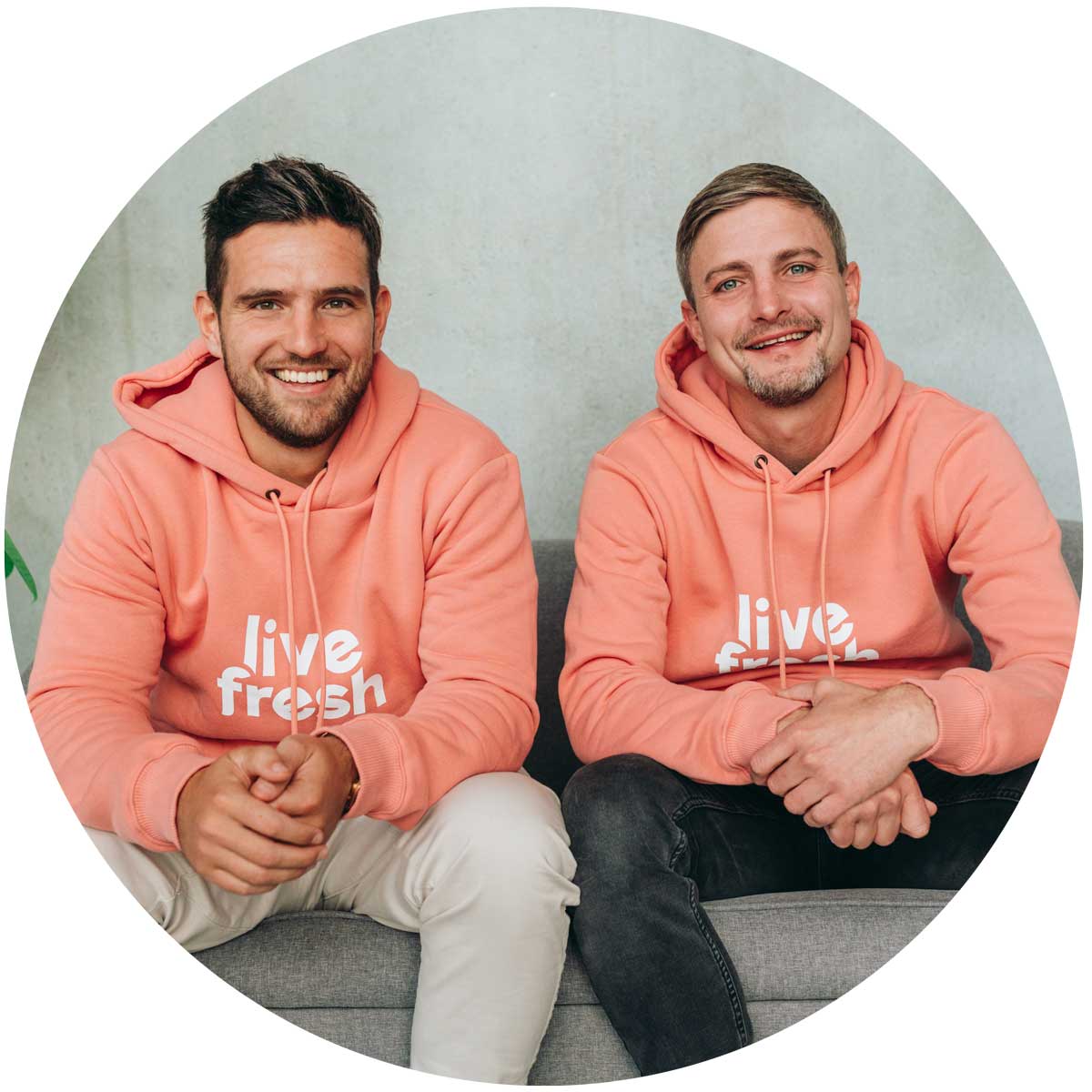 Das Bild, in einem Kreis dargestellt, zeigt zwei Männer in korallenfarbenen Hoodies mit dem Logo "Live Fresh", die entspannt auf einem grauen Sofa sitzen. Sie lächeln freundlich in die Kamera, was eine sympathische und zugängliche Atmosphäre vermittelt.