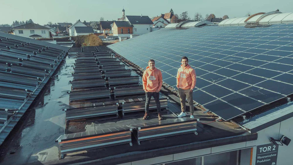 Zwei Männer, die auf einem Dach voller Solarmodule stehen. Beide tragen orangefarbene Hoodies mit dem „live fresh“-Logo. Im Hintergrund ist eine ländliche Umgebung mit Häusern und Bäumen zu sehen. Das Bild vermittelt das Engagement des Unternehmens für Nachhaltigkeit und Klimaneutralität durch die Nutzung erneuerbarer Energien, was durch die Solaranlagen auf dem Dach unterstrichen wird. Die Szene wirkt zukunftsorientiert und ökologisch bewusst.