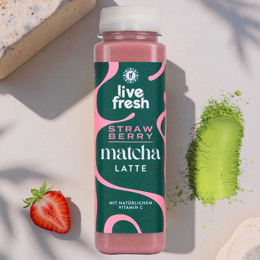 Eine Flasche Strawberry Matcha Latte von LiveFresh auf hellem Hintergrund. Daneben eine aufgeschnittene Erdbeere und Matchapulver.