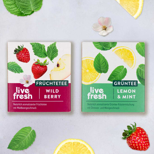 Zwei Teepackungen von LiveFresh – links „Früchtetee Wild Berry“ mit Erdbeeren und Apfel, rechts „Grüntee Lemon & Mint“ mit Zitronenscheiben und Minzblättern; dekoriert mit passenden Früchten und Blättern.