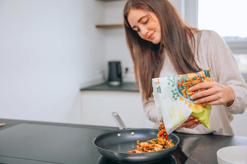 Eine Frau in einer modernen Küche, die eine „Live Fresh“-Verpackung mit der Aufschrift „Bowl Exotic Style“ in eine Pfanne gibt. In der Pfanne befinden sich bereits bunte Gemüsestücke, die als Teil eines gesunden, veganen Gerichts zubereitet werden. Die Frau lächelt leicht und scheint konzentriert bei der Zubereitung zu sein.