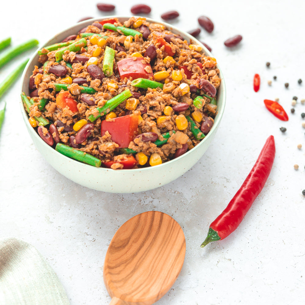 Eine Schale mit "Chili sin Carne", einer veganen Mahlzeit. In der Schale sind grüne Bohnen, Kidneybohnen, Mais, Tomaten und pflanzliches Hack zu sehen. Um die Schale herum liegen frische Zutaten wie Chilischoten und Pfefferkörner, was die würzige Natur des Gerichts unterstreicht.