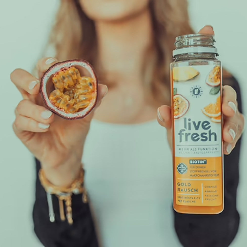 Eine Frau hält in einer Hand links vom Bild eine aufgeschnittene Passionsfrucht und in der anderen eine geöffnete Flasche "Gold Rausch" Saft von LiveFresh. Das Etikett zeigt Bilder von Orangen, Ananas und Passionsfrucht.