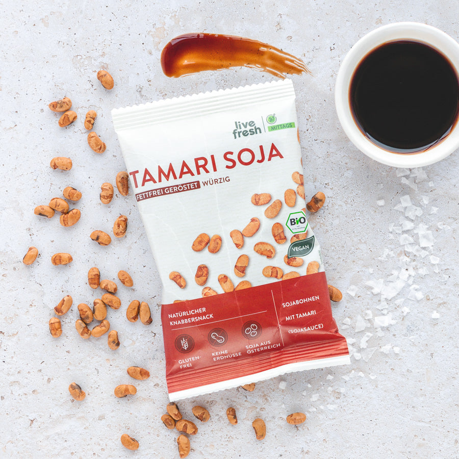 Packung 'Live Fresh Tamari Soja' mit gerösteten Sojabohnen und einem Klecks dunkler Sojasauce daneben, präsentiert auf einem hellen, steinernen Hintergrund.