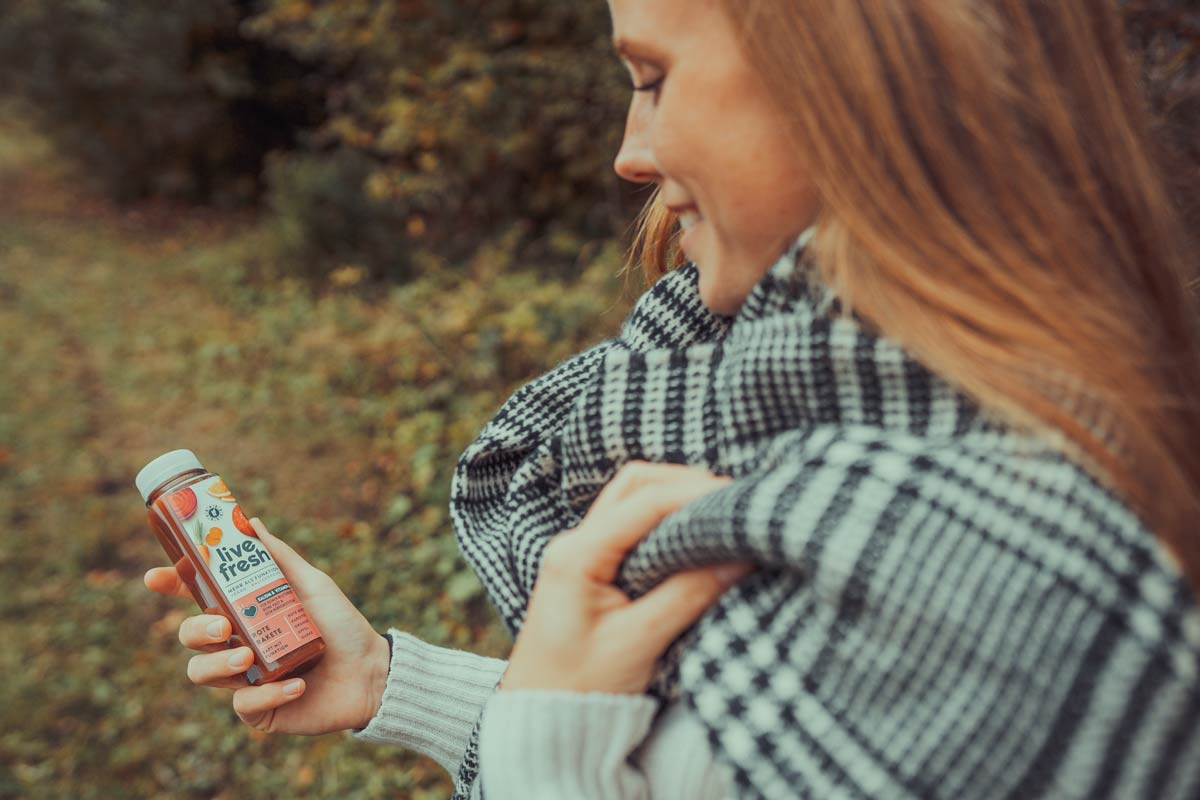 Eine Frau im Freien, die eine Flasche LiveFresh Rote Rakete Saft in der Hand hält. Sie ist in einen schwarz-weiß karierten Schal gehüllt, während sie lächelnd die Saftflasche betrachtet. Die herbstliche Umgebung mit Bäumen und Laub verleiht dem Bild eine gemütliche Atmosphäre.
