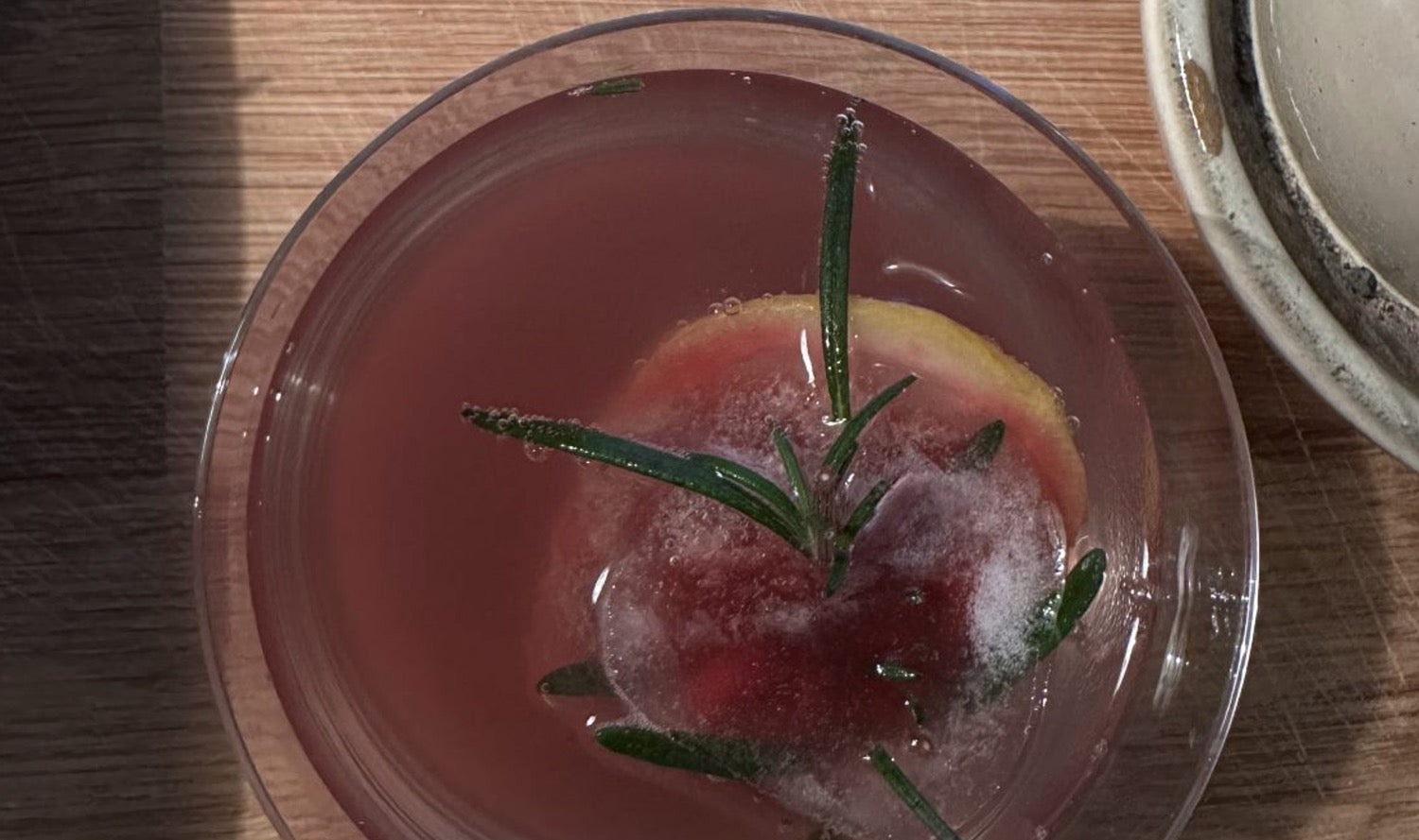 Ein Silvester-Cocktail mit klarem, rosa gefärbtem Getränk, garniert mit einem Rosmarinzweig und einer Zitronenscheibe im Eis, serviert in einem eleganten Glas auf einem Holztisch.