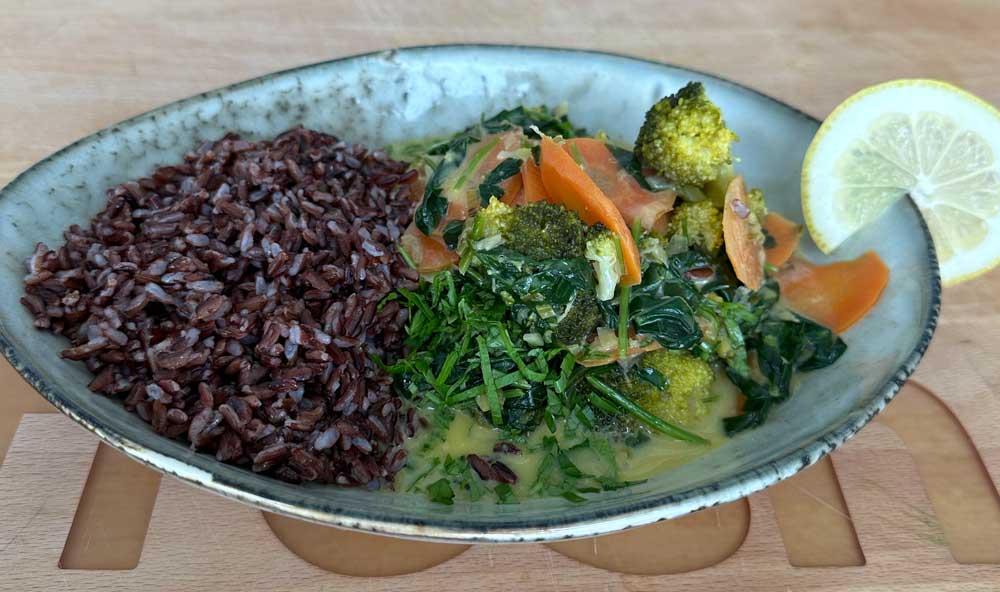 Eine Portion Curry mit vielfältigem Gemüse in Kokosmilch und frischen Kräutern an der Seite, begleitet von einer Portion dunklem Reis und einer Zitronenscheibe auf einem rustikalen Teller.