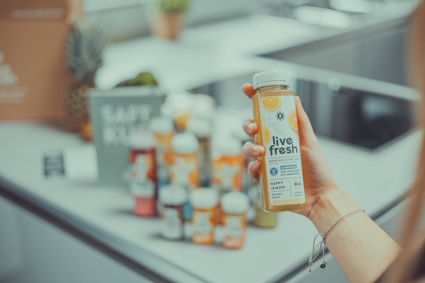Eine Hand hält ein LiveFresh Saft namens Happy Lemon in der Hand. Im Hintergrund sind verschwommen weitere Säfte und Shots zu sehen, sowie ein Saftkurguide, eine Ananas und ein Karton auf heller Arbeitsfläche.