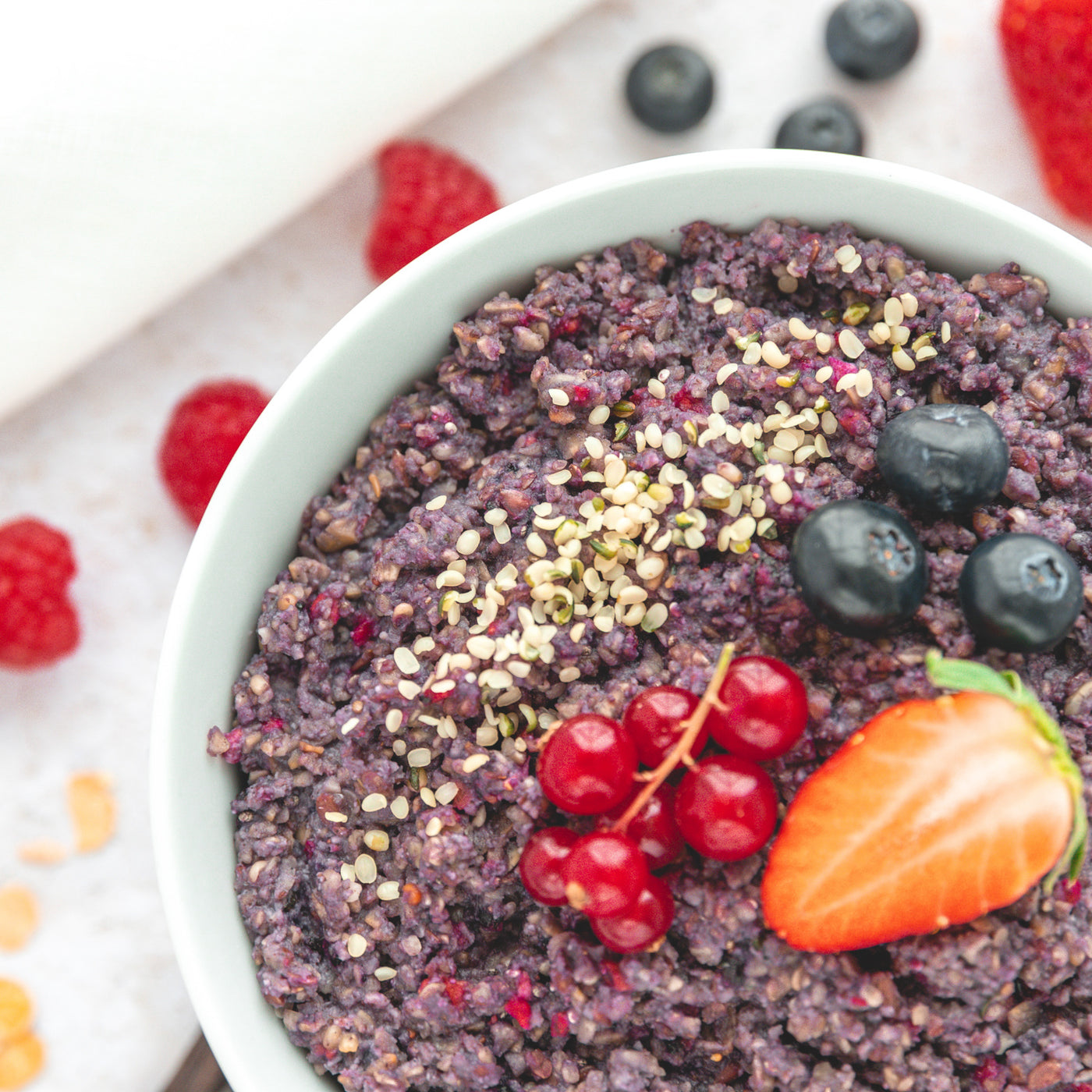 Ausschnitt einer weißen Schüssel mit Chiapudding, garniert mit Stachelbeeren, Heidelbeeren und Erdbeeren, welches auf den Beerengeschmack des Puddings hinweist. Im hellen Hintergrund sind Himbeeren und Heidelbeeren zu sehen.