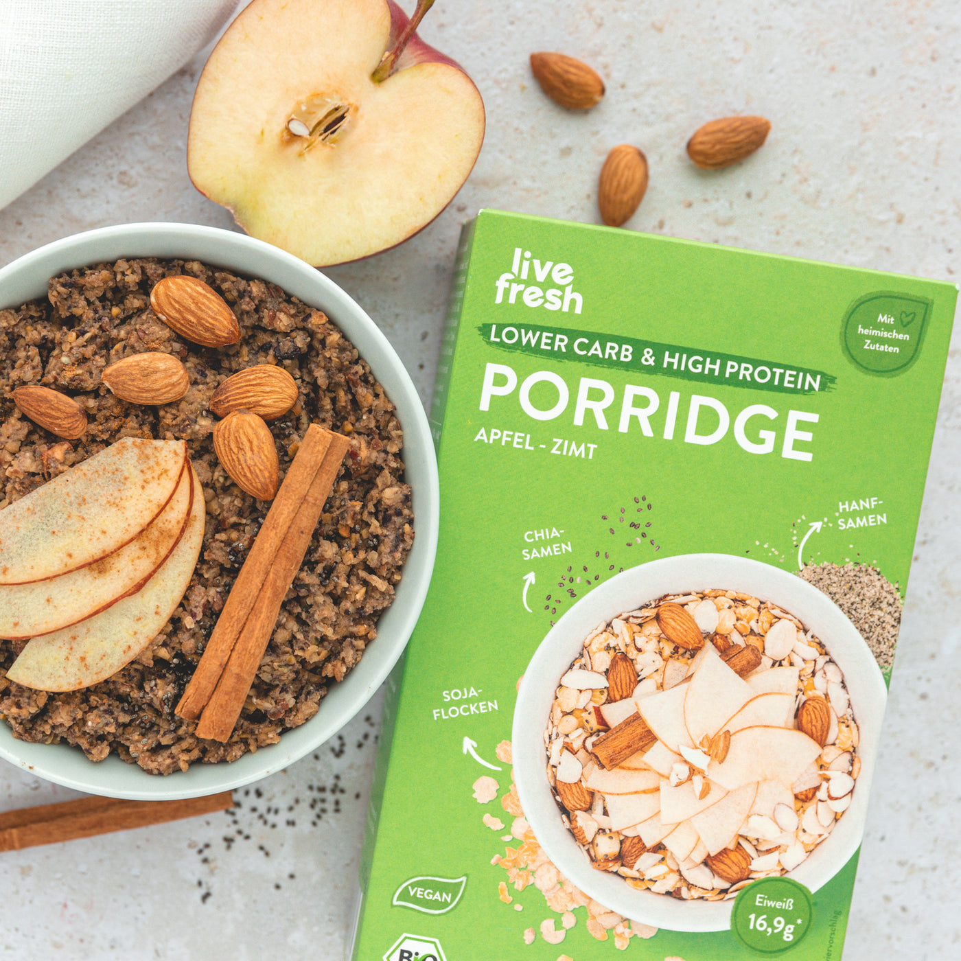 Eine Schüssel Apfel-Zimt Porridge von LiveFresh, garniert mit Apfelscheiben, Mandeln und Zimtstangen. Daneben die weiß-grüne Verpackung mit dem Hinweis „Lower Carb & High Protein“, vegan und aus heimischen Zutaten wie Sojaflocken, Chiasamen und Hanfsamen, mit 17g Eiweiß pro Portion. Daneben ein halbierter Apfel, Mandeln und Zimtstangen.