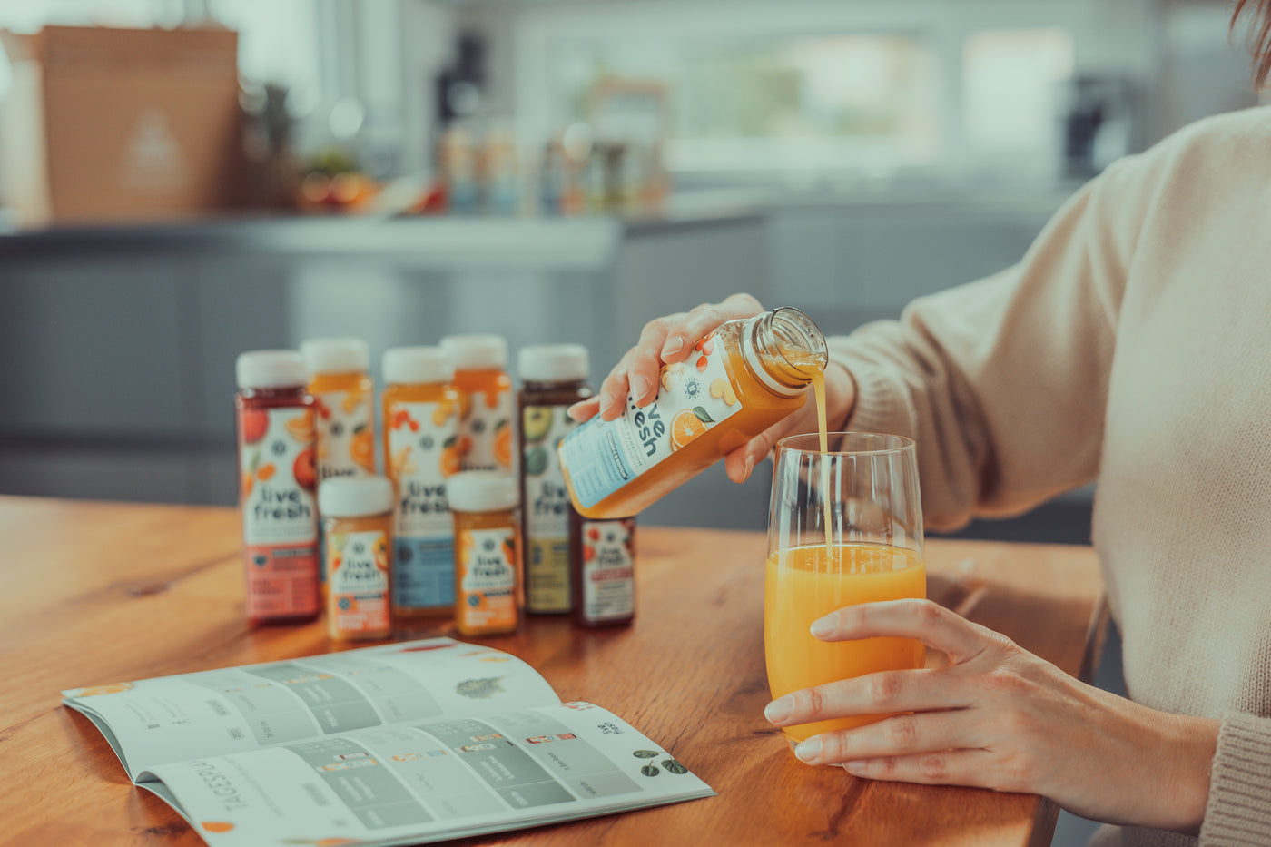 Person gießt einen gelben LiveFresh-Saft in ein Glas, im Vordergrund liegt ein geöffneter Saftkur-Guide, im Hintergrund stehen mehrere Saftflaschen auf einem Holztisch.