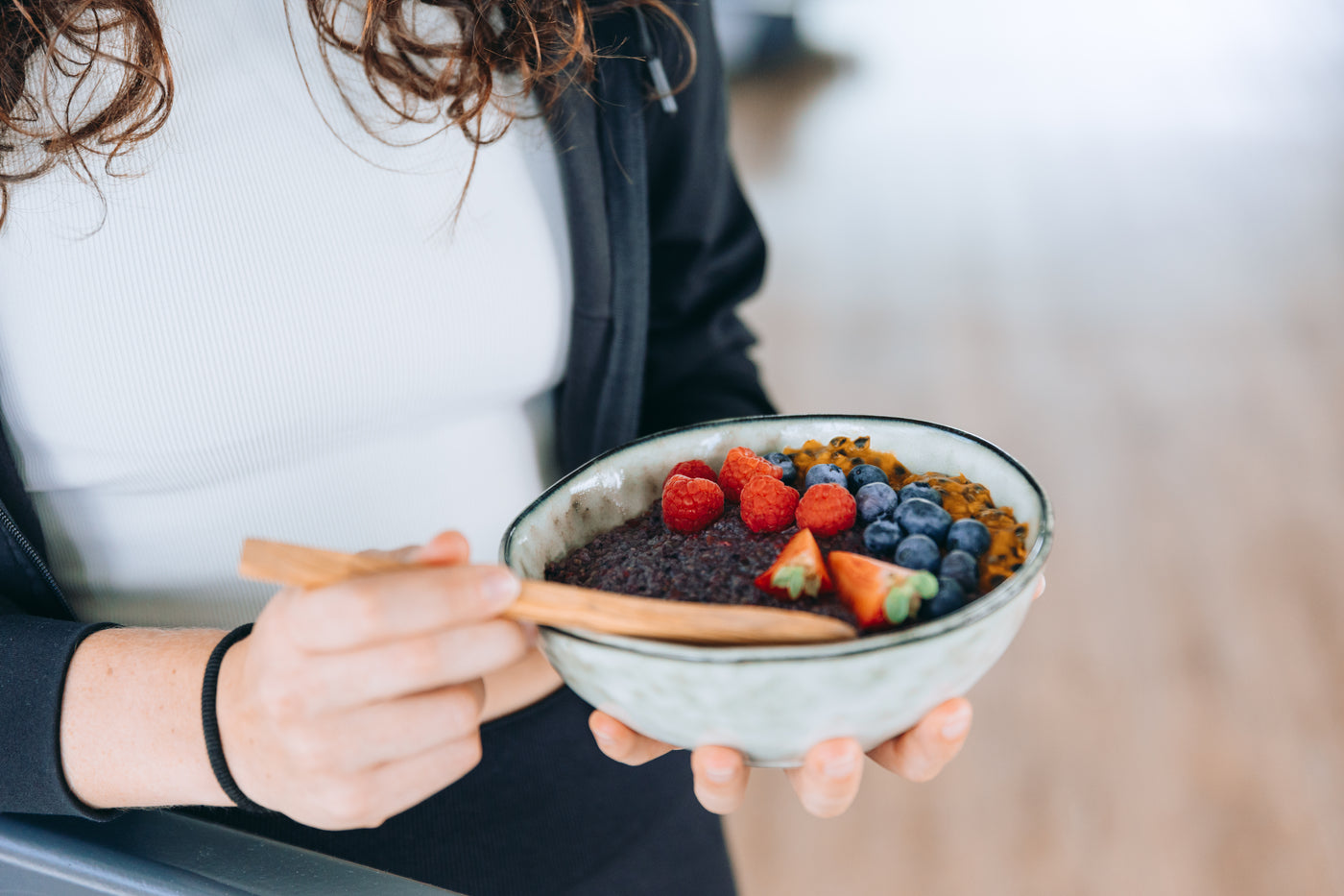 Person hält eine Schüssel mit einem bunten Frühstück aus Beeren, Chia-Pudding und Passionsfrucht, mit einem Holzlöffel in der Hand.