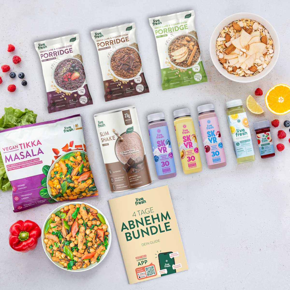 Flatlay verschiedener LiveFresh Produkte auf hellem Hintergrund mit Porridge Beuteln, Slim Shake, SKVR Protein Drinks, Saft Shots und veganer Tikka Masala Mahlzeit neben frischem Obst und einer Schüssel Essen. In der Mitte liegt ein Heft mit der Aufschrift 4 Tage Abnehm Bundle Dein Guide.