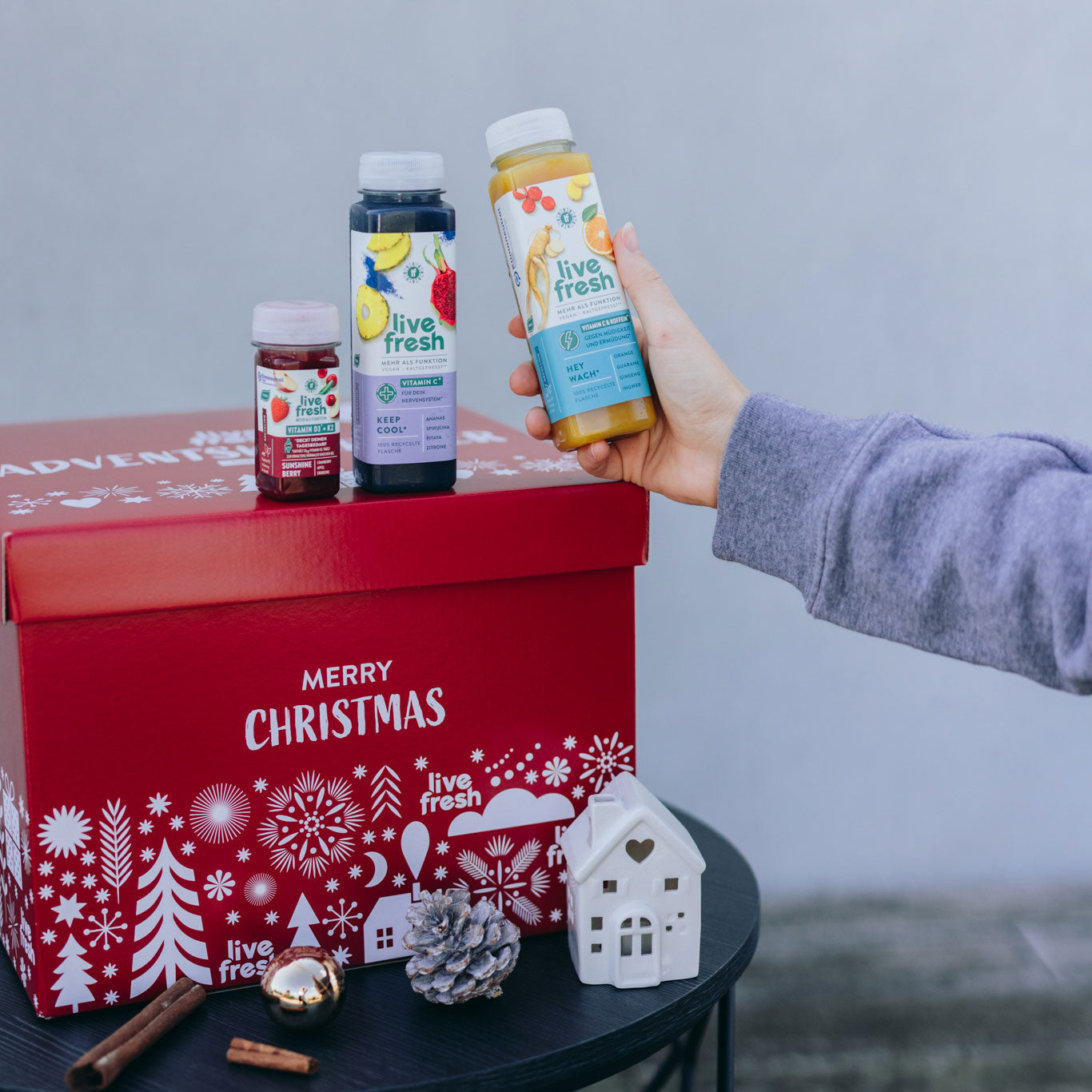 Eine rote "Merry Christmas" Box von live fresh steht auf einem Tisch, dekoriert mit Tannenzapfen und weihnachtlichen Elementen. Darauf stehen drei verschiedene Flaschen von live fresh Säften, während eine Hand eine Flasche hält.