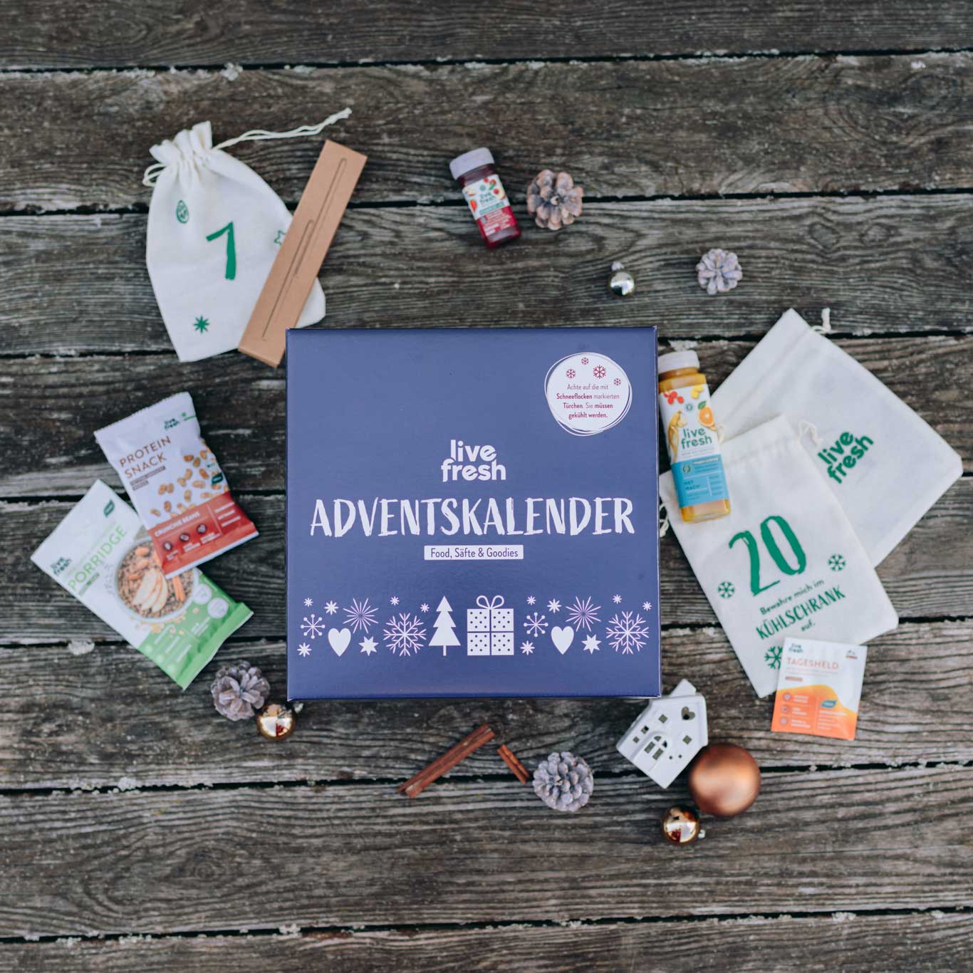 Live Fresh Adventskalender-Box mit dem Schriftzug "Food, Säfte & Goodies" steht auf einem rustikalen Holztisch. Um die Box herum liegen verschiedene Überraschungen, wie Protein-Snacks, kleine Säfte, dekorative Tannenzapfen und nummerierte Stoffbeutel, die den Inhalt des Kalenders andeuten.