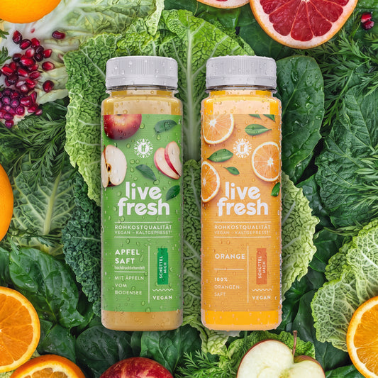 Zwei gekühlte Flaschen Live Fresh Apfel- und Orangensaft stehen auf einem Bett aus frischem Blattgemüse, umgeben von Äpfeln, Orangen, Grapefruit und Granatapfel. Die frischen Zutaten vermitteln die Vielfalt eines Frühstückspakets mit kaltgepressten Säften für einen ausgewogenen Start in den Tag.