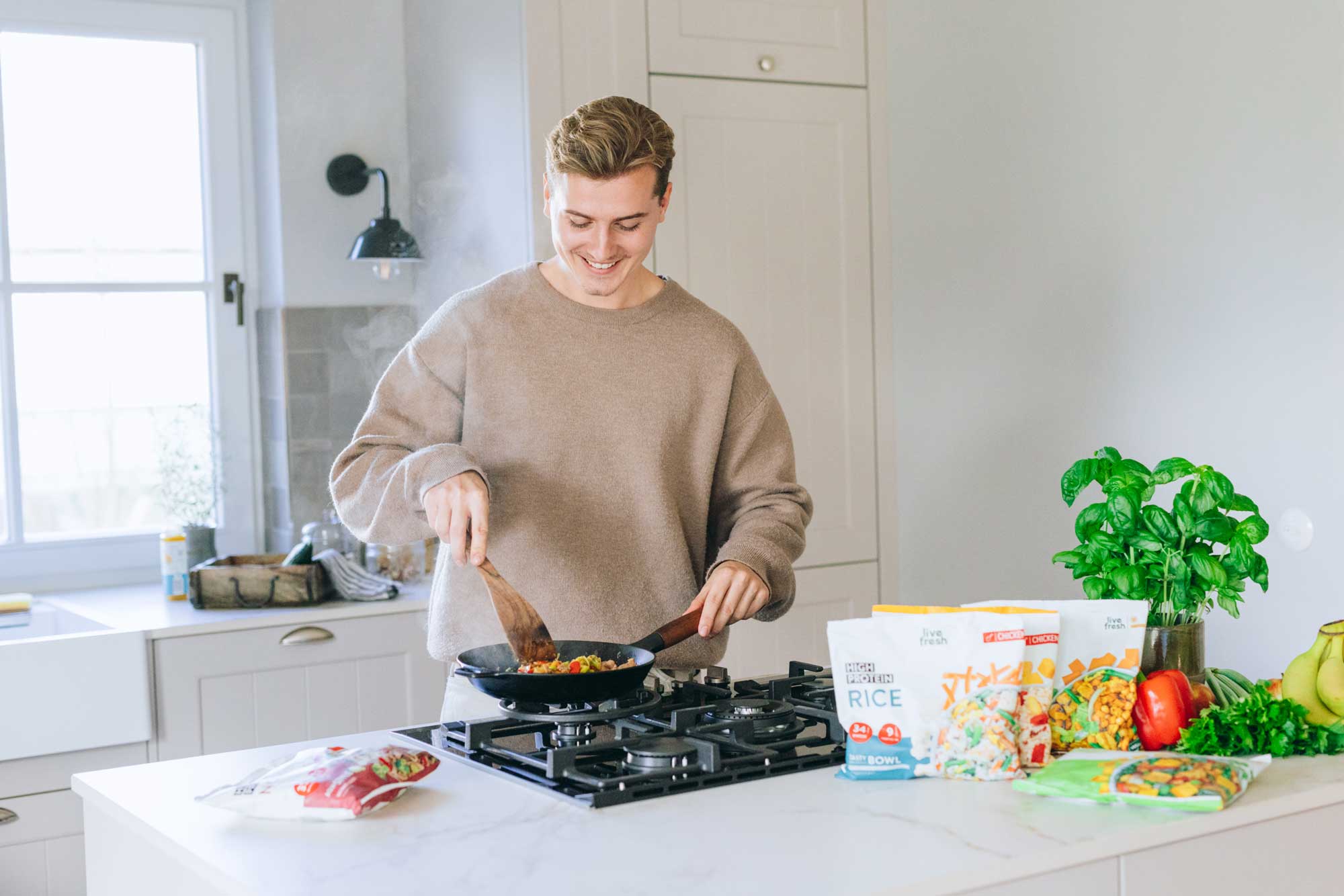 Junger Mann kocht in einer modernen Küche mit einer Pfanne auf dem Herd, umgeben von LiveFresh Tiefkühlgerichten, frischem Gemüse und Kräutern auf der Arbeitsfläche.