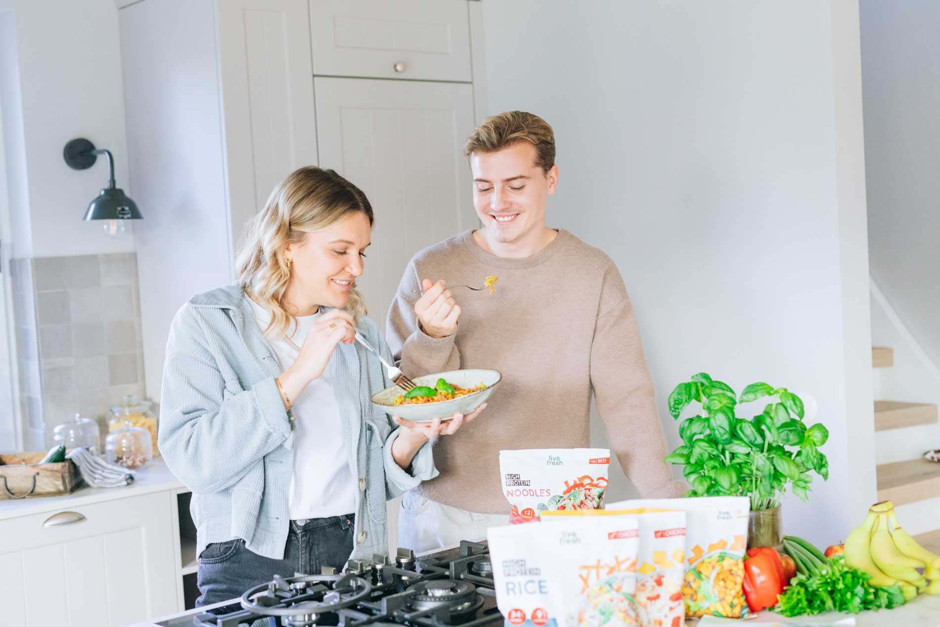 Junges Paar steht in einer hellen Küche und genießt gemeinsam eine Bowl mit LiveFresh Tiefkühlgericht; im Vordergrund frisches Gemüse und mehrere Produktpackungen.