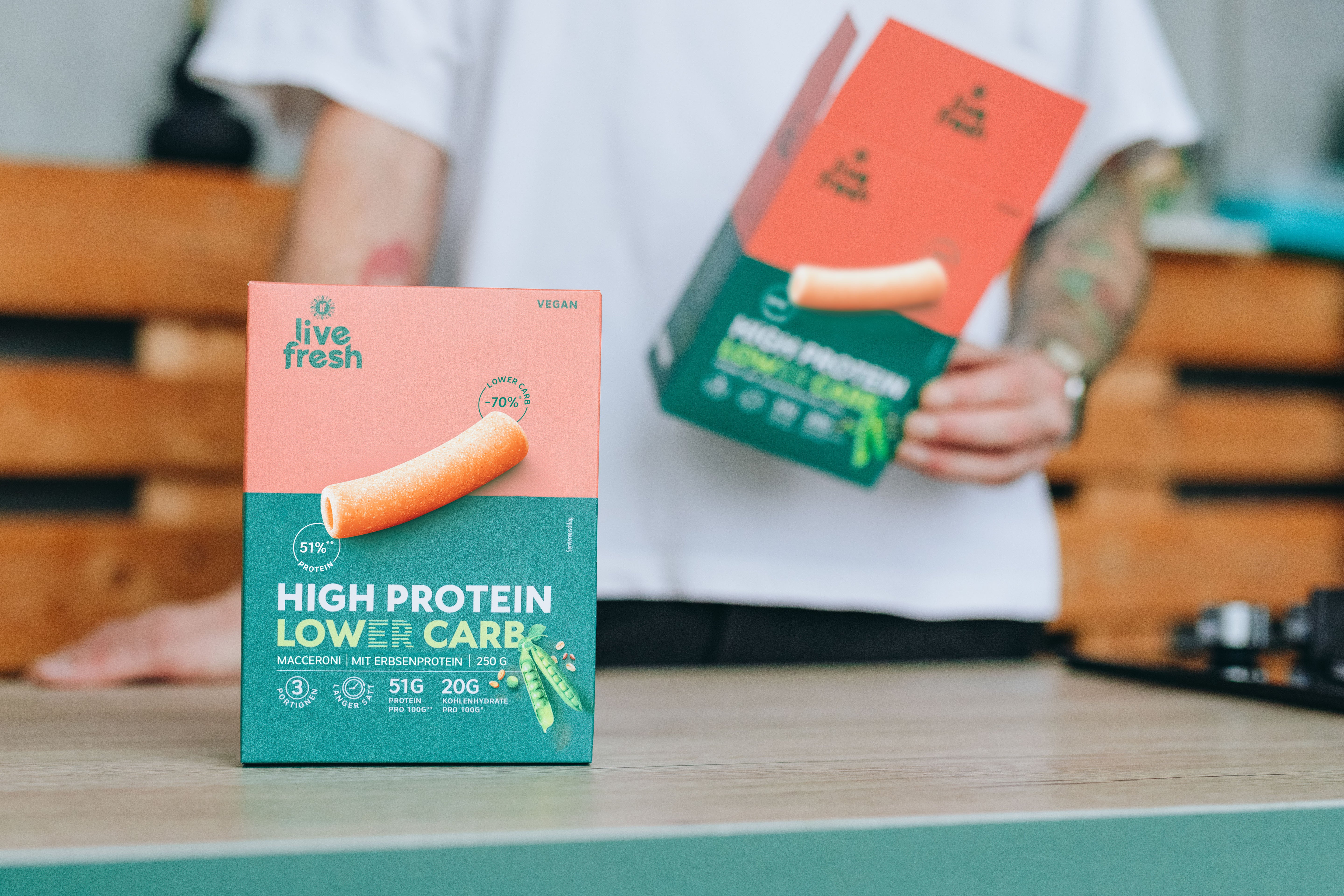 Verpackung von Protein Pasta auf einer Küchenarbeitsplatte. Vorn steht eine Box mit der Aufschrift „live fresh“, „VEGAN“ und „HIGH PROTEIN LOWER CARBS“, während im unscharfen Hintergrund eine Person eine zweite Packung hält.