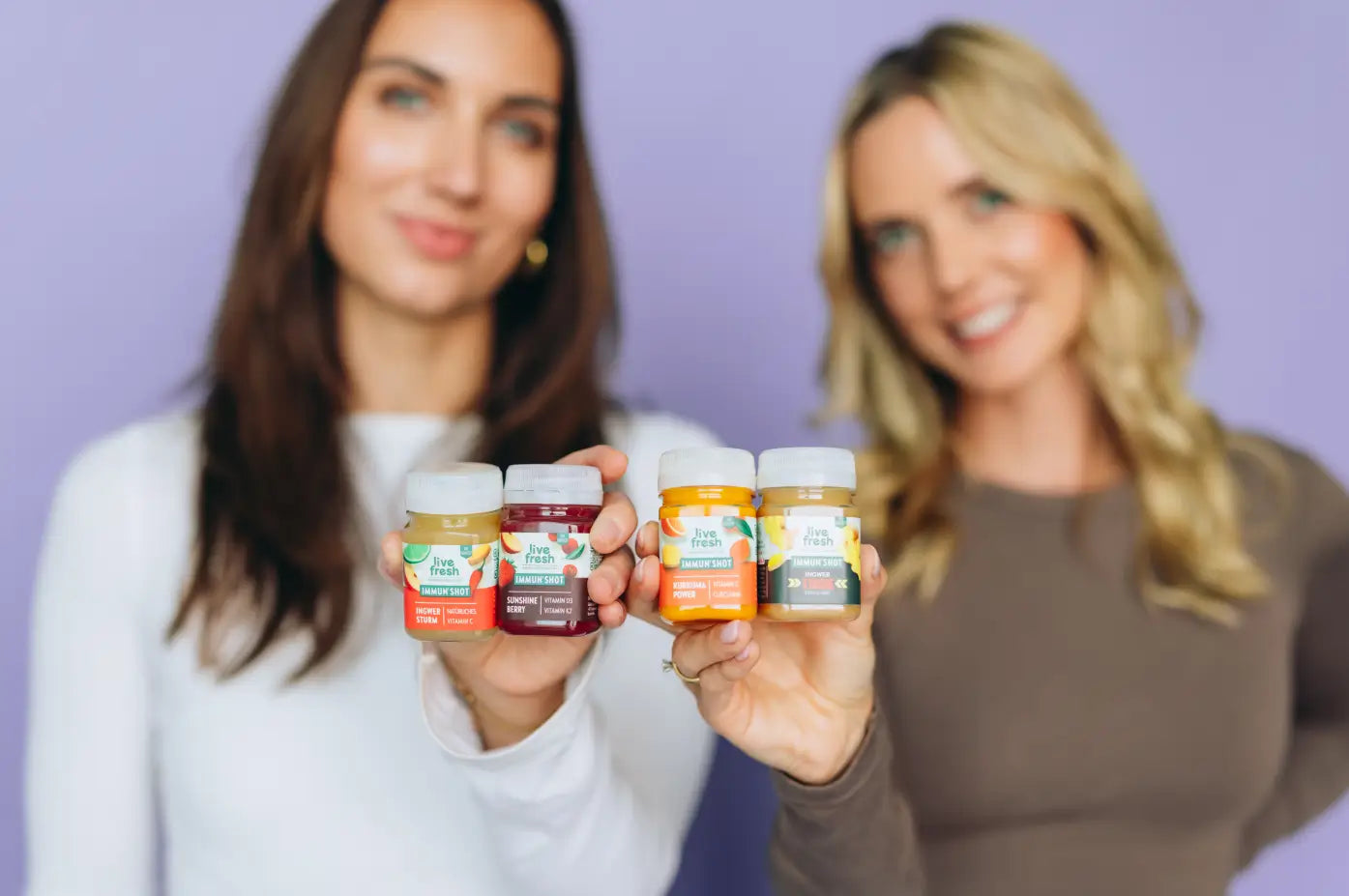 Zwei lächelnde Frauen halten vier LiveFresh Immunshots in die Kamera. Die Sorten Ingwer Sturm mit Vitamin C. Sunshine Berry mit Vitamin D3 und K2. Kurkuma Power mit Vitamin C und Curcumin sowie Ingwer Xtreme Extra Scharf sind deutlich zu erkennen.