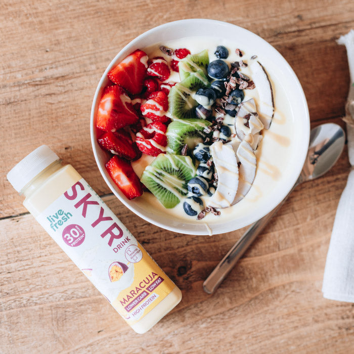 Eine Bowl, garniert mit Erdbeeren, Himbeeren, Kiwi, Blaubeeren und Kakaonibs. Daneben steht eine Flasche „Live Fresh Skyr Drink Maracuja“ mit 30g Protein. Die Skyr-Flasche wirbt mit niedrigem Kohlenhydratgehalt, fettarm und proteinreich, und betont eine gesunde und ausgewogene Ernährungswahl.