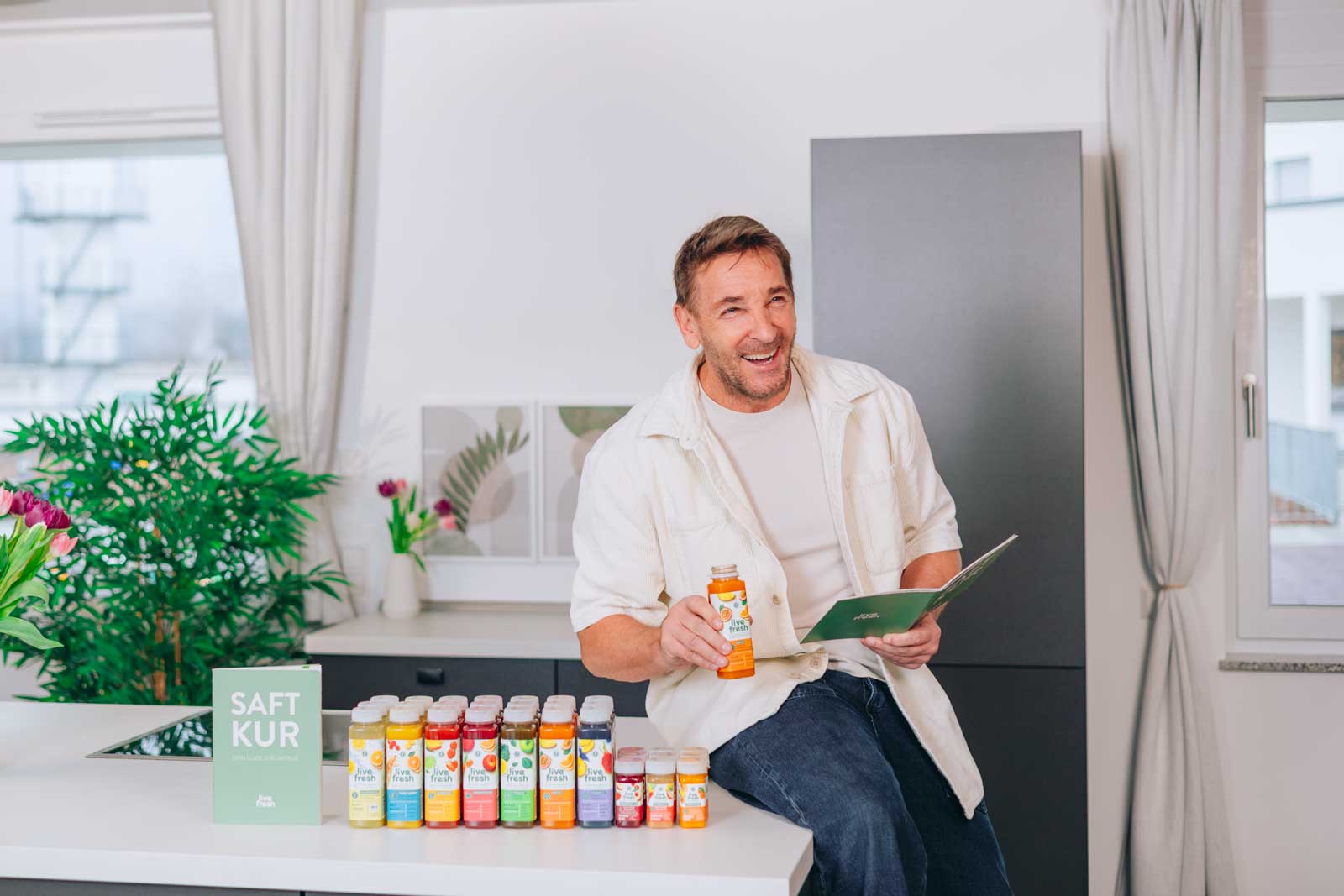 Ein lachender Mann sitzt in einer modernen Küche, hält eine Saftflasche und ein grünes Heft. Vor ihm stehen bunte Säfte und ein Schild mit „SAFT KUR“. Im Hintergrund sind Pflanzen und Deko.