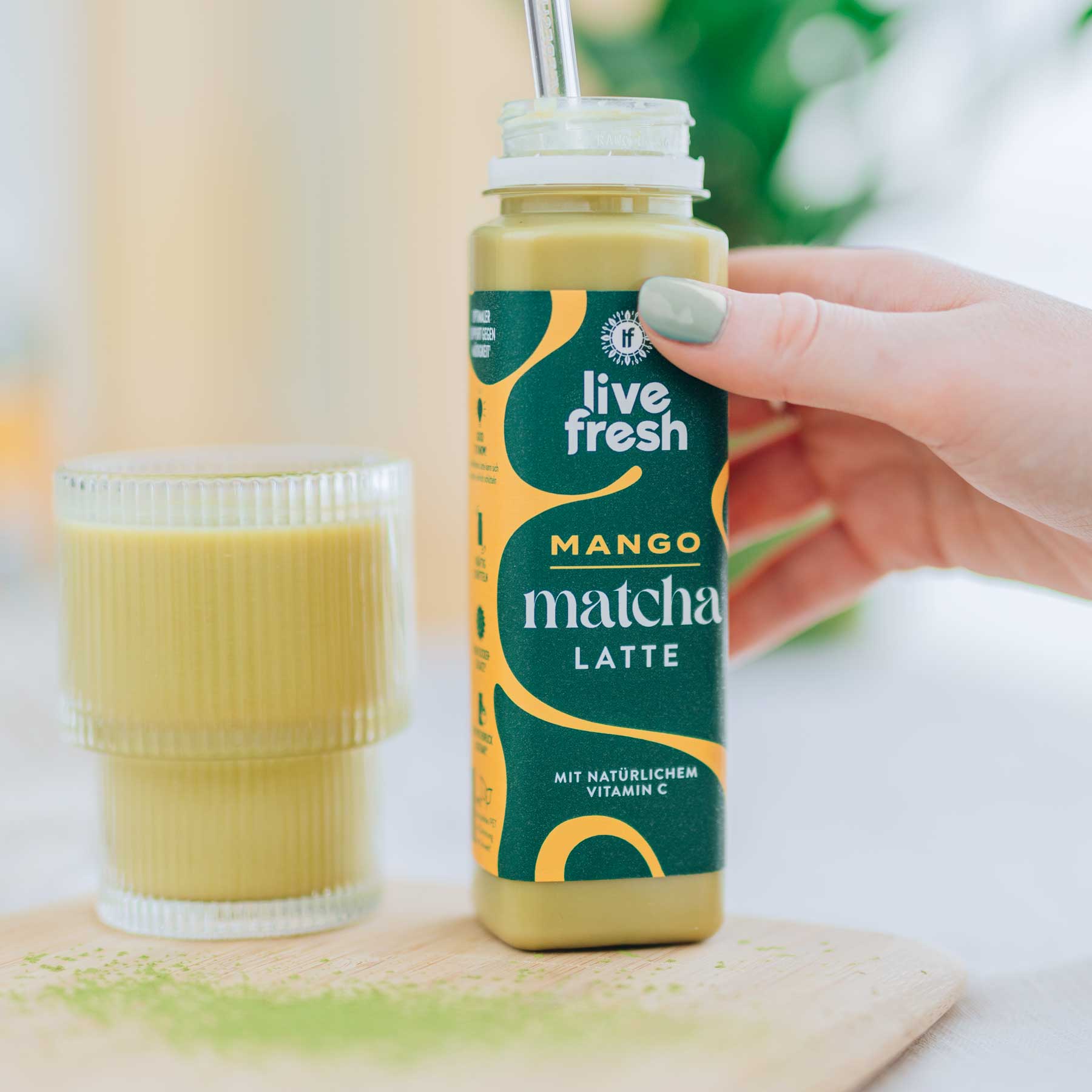 Hand hält „Live Fresh“ Mango Matcha Latte mit Strohhalm, daneben gefülltes Glas auf Holzbrett, im Hintergrund weiches Tageslicht.
