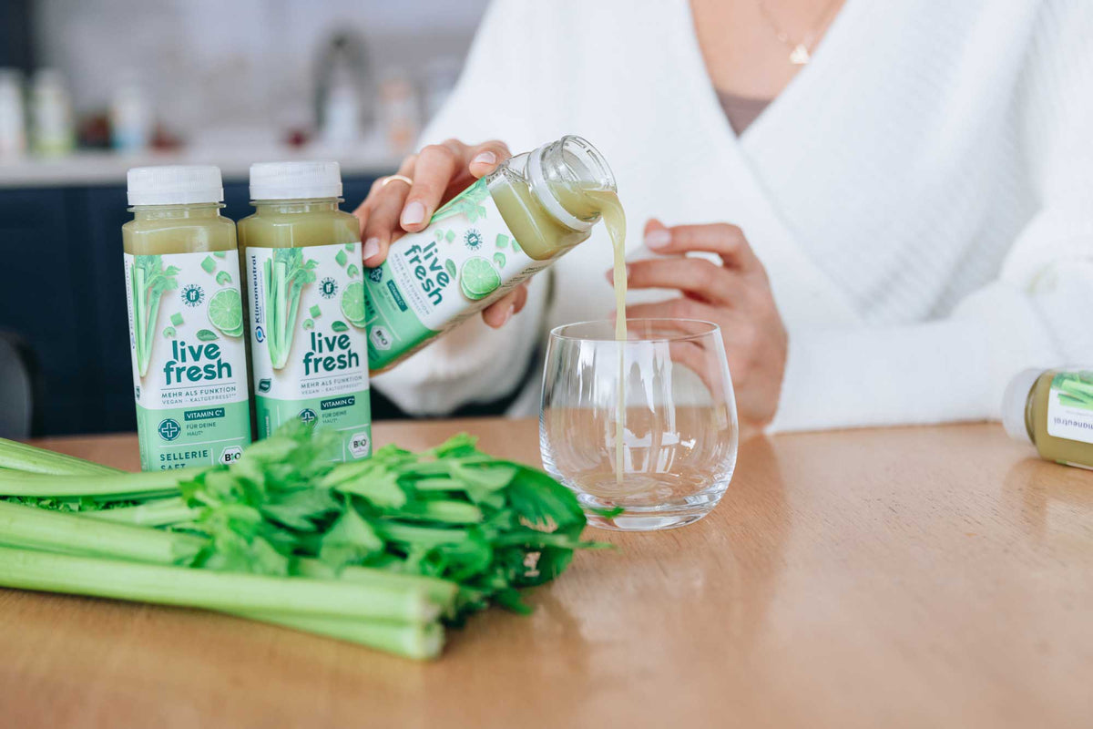 Frau gießt LiveFresh BIO Selleriesaft aus der Flasche in ein Glas auf der Küchentheke mit frischen Selleriestangen und weiteren Selleriesaft-Flaschen – LiveFresh