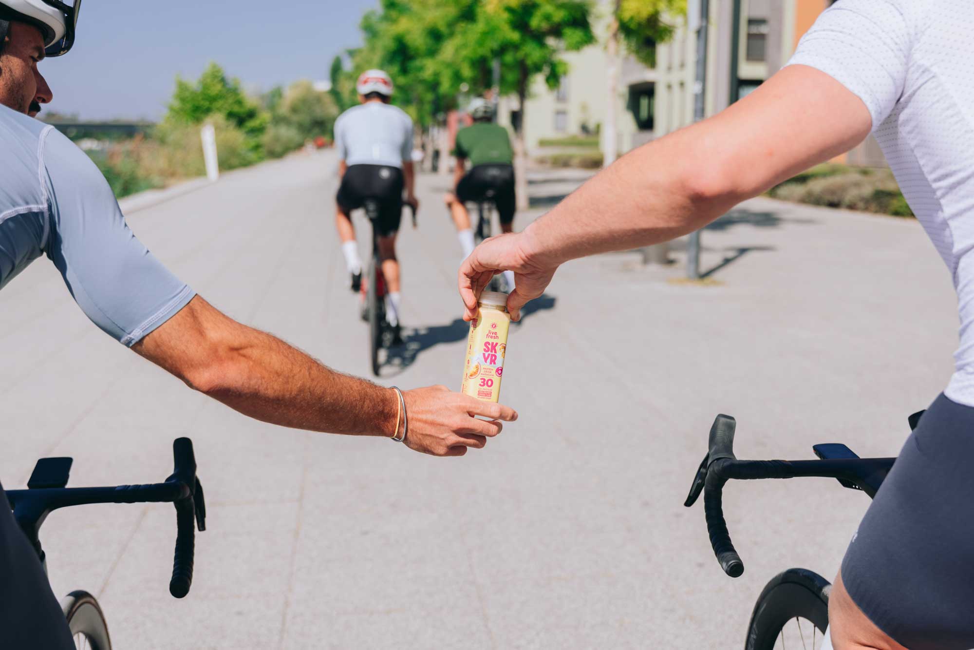 Zwei Radfahrer im Trikot übergeben sich während der Fahrt eine Flasche Skyr-Drink von LiveFresh auf einem sonnigen Stadtweg mit Bäumen im Hintergrund.