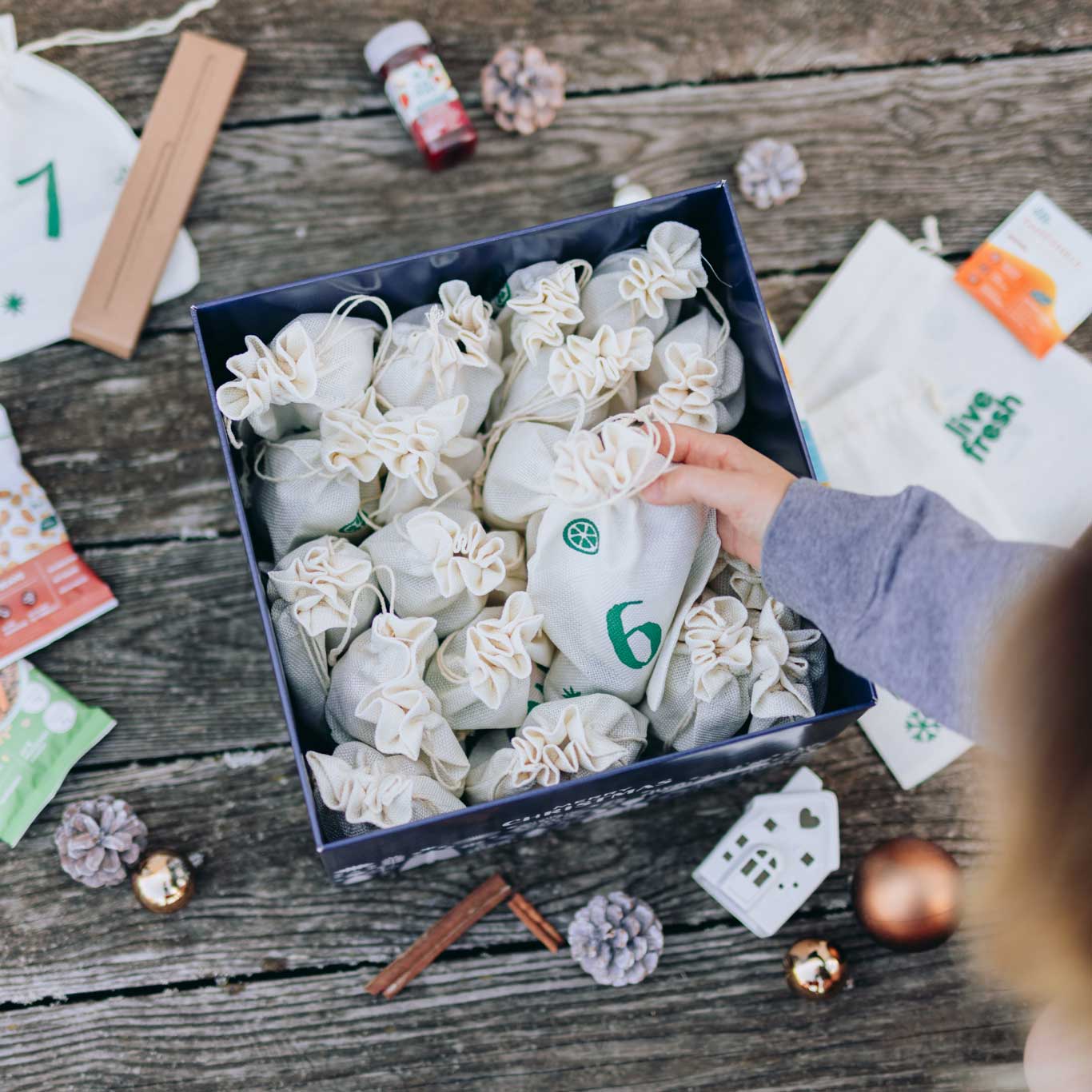 Live Fresh Adventskalender-Box mit dem Schriftzug "Food, Säfte & Goodies" steht auf einem rustikalen Holztisch. Um die Box herum liegen verschiedene Überraschungen, wie Protein-Snacks, kleine Säfte, dekorative Tannenzapfen und nummerierte Stoffbeutel, die den Inhalt des Kalenders andeuten.