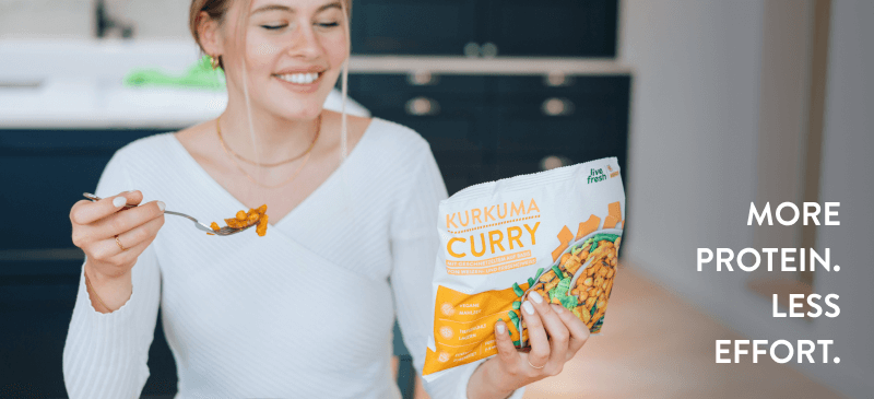 Woman with white sweater is eating while she looks at a package of a ready-to-eat meal named 