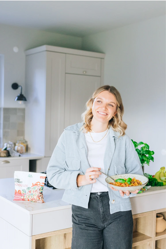Frau steht in moderner Küche mit einer Bowl in der Hand und lächelt, im Vordergrund eine LiveFresh Fertiggerichte High Protein Nudelpackung