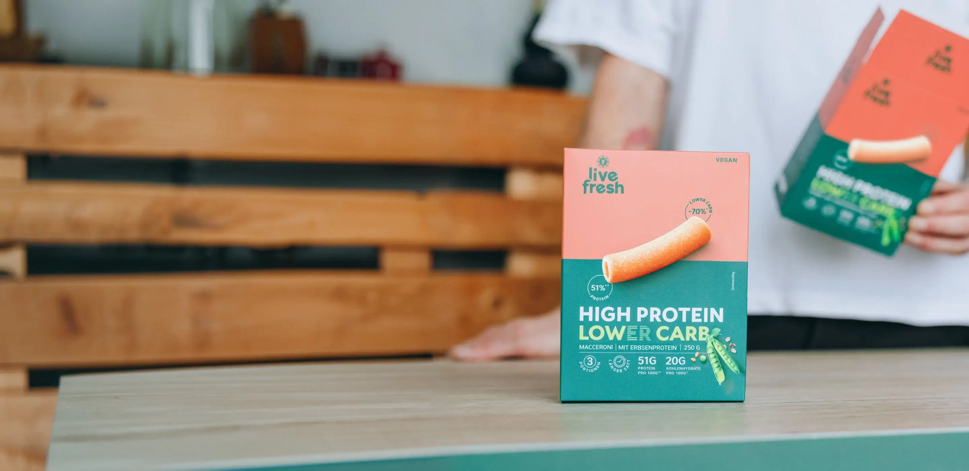 Packung LiveFresh High Protein Lower Carb Maccharoni aus Erbsenprotein steht auf einer Küchenarbeitsplatte. Eine Person hält im Hintergrund eine weitere Pasta Verpackung.