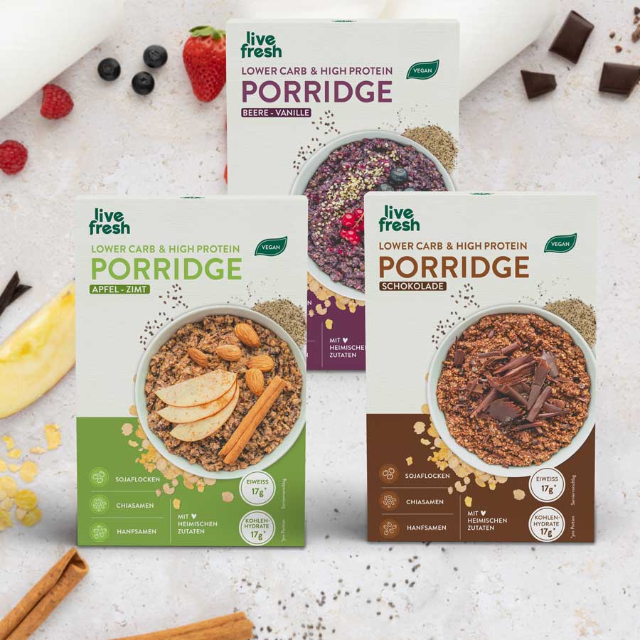 Porridge-Probierpaket: Drei Packungen "Live Fresh Lower Carb & High Protein Porridge" in den Sorten Apfel-Zimt, Beere-Vanille und Schokolade stehen nebeneinander. Auf dem Tisch liegen Zutaten wie Beeren, Zimtstangen, Chiasamen und Schokoladenstücke.