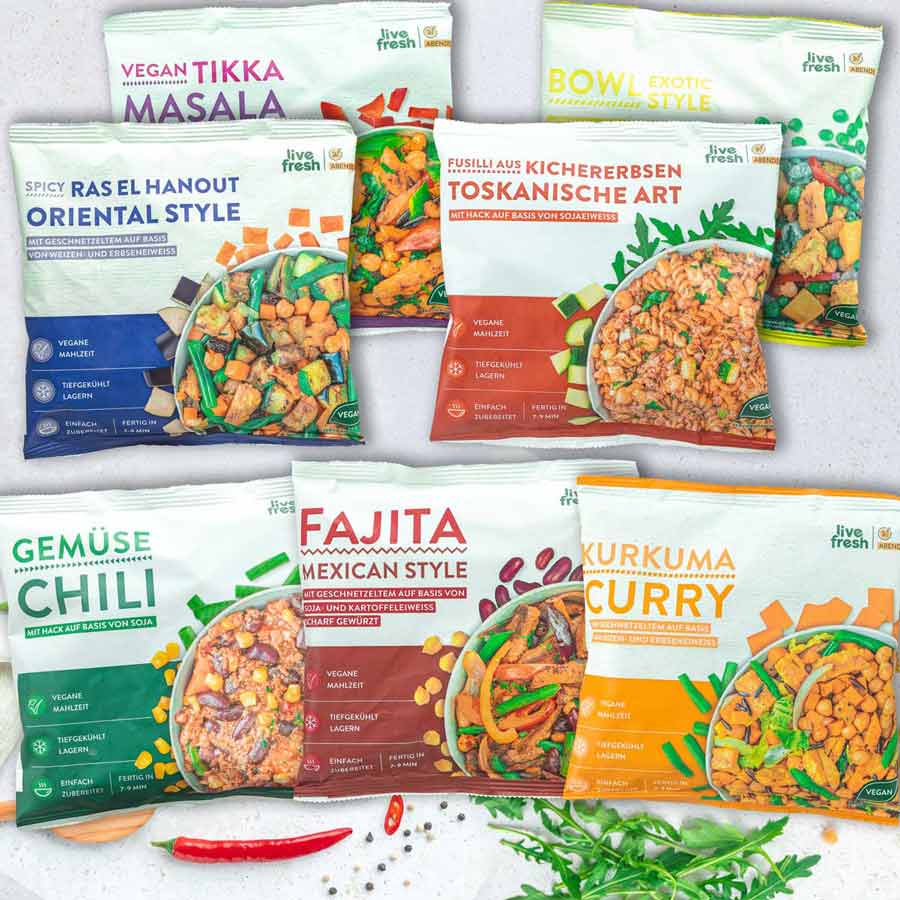 Sieben vegane Tiefkühlgerichte von LiveFresh auf einer hellen Oberfläche. Enthalten sind Vegan Tikka Masala, Bowl Exotic Style, Spicy Ras El Hanout Oriental Style, Fusilli aus Kichererbsen – Toskanische Art, Gemüse Chili, Fajita Mexican Style und Kurkuma Curry. Die Verpackungen zeigen die jeweiligen Gerichte in verschiedenen Farben mit buntem Gemüse und pflanzlichem Protein. Rundherum liegen Zutaten wie Chili, Kräuter und Gewürze.