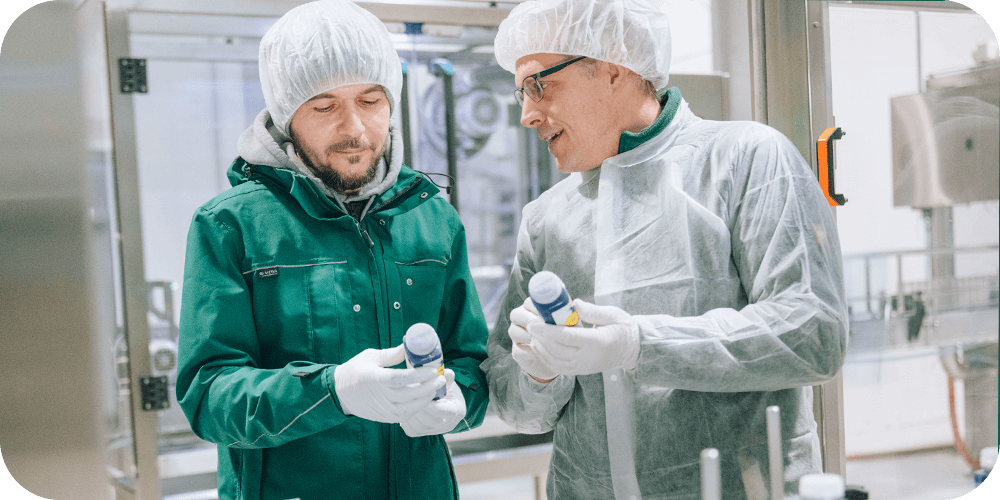 Zwei Männer in Hygienekleidung mit Haarnetz, Handschuhen und Schutzkittel stehen in einer Produktionshalle und begutachten jeweils eine LiveFresh Saftflasche. Einer trägt zusätzlich eine grüne Jacke. Im Hintergrund sind industrielle Maschinen zu sehen, was auf einen kontrollierten und professionellen Herstellungsprozess hinweist.





