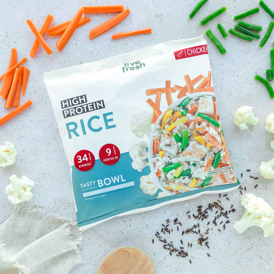 Das Bild zeigt eine Packung „Live Fresh High Protein Rice“ Tasty Bowl mit Hähnchenbrustfilet. Die Verpackung hebt einen Proteingehalt von 34 g und eine Zubereitungszeit von 9 Minuten hervor. Rund um die Packung liegen frische Zutaten wie Karotten, grüne Bohnen und Blumenkohl, was die ausgewogene und proteinreiche Mahlzeit betont.