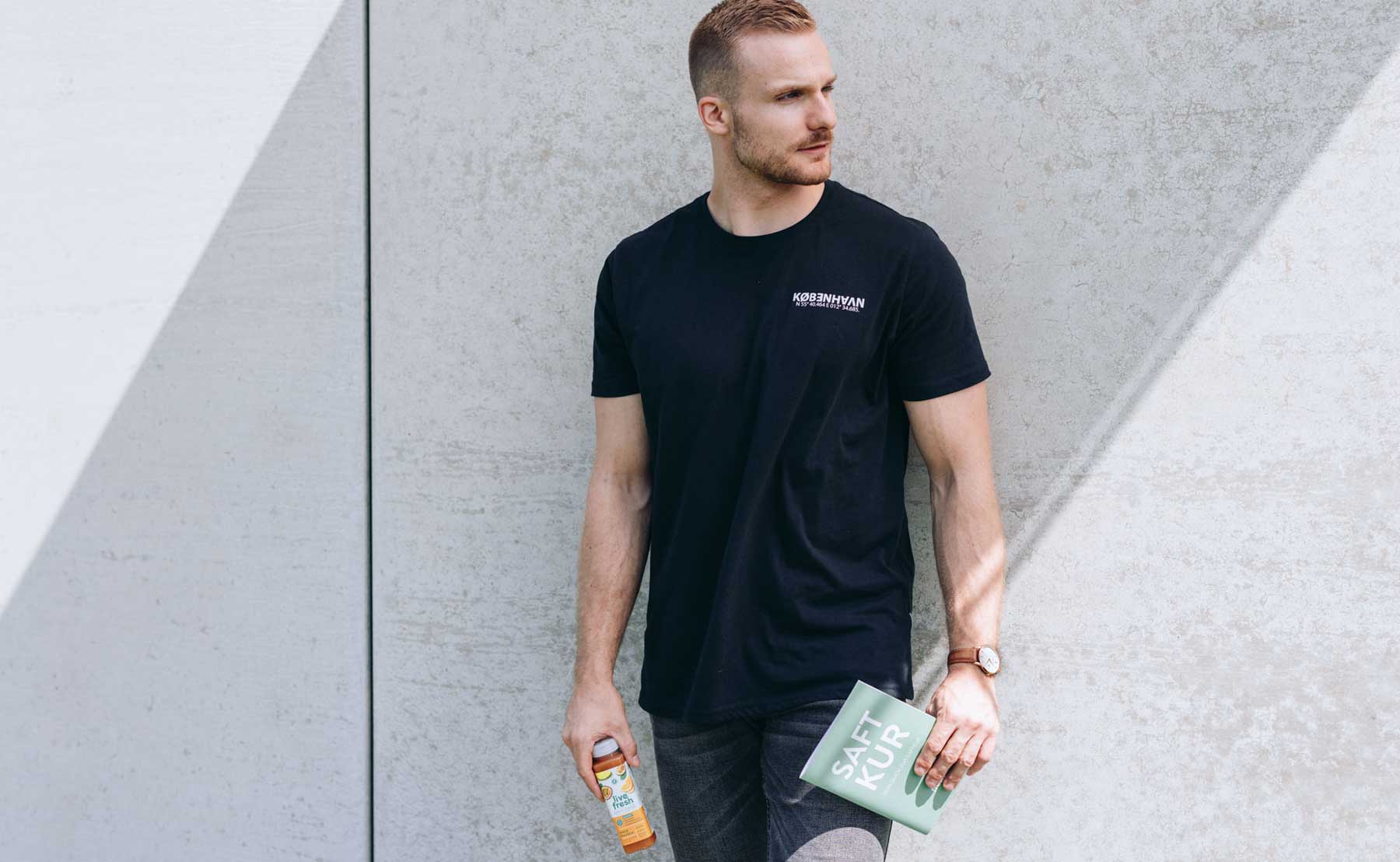 Ein sportlicher Mann lehnt an einer Betonwand und hält in einer Hand eine grüne Broschüre mit der Aufschrift „SAFT KUR“ und in der anderen eine LiveFresh Saftflasche. Er trägt ein schwarzes T-Shirt und eine Armbanduhr, während Sonnenlicht geometrische Schatten auf die Wand wirft.