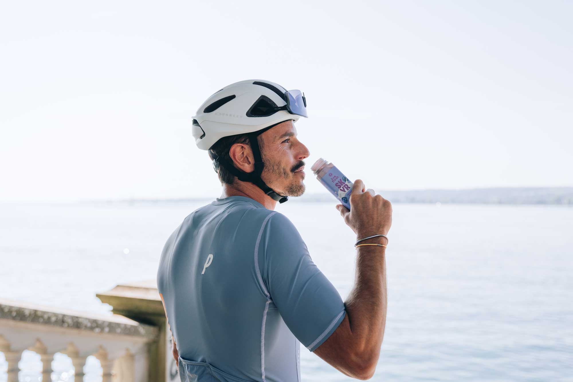 Radfahrer mit weißem Helm und blaugrauem Trikot trinkt ein Skyr-Drink von LiveFresh mit Blick aufs Wasser bei hellem Tageslicht.